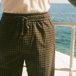 A close-up of a person wearing dark brown and black checkered drawstring shorts, standing on a deck with the blue ocean and white railing in the background.