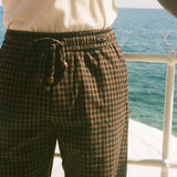 A close-up of a person wearing dark brown and black checkered drawstring shorts, standing on a deck with the blue ocean and white railing in the background.
