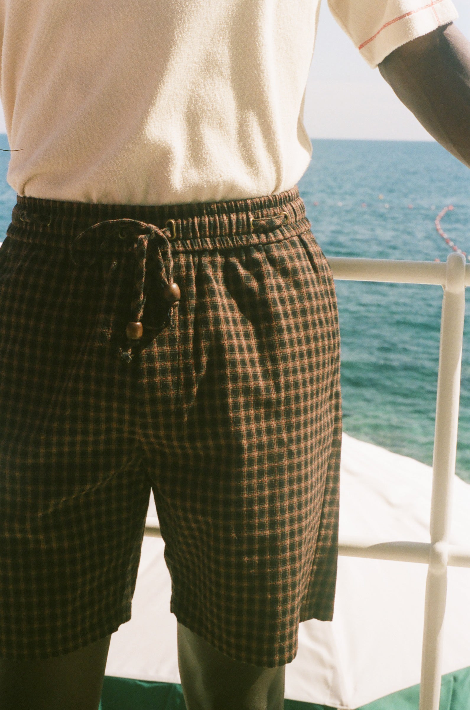 A close-up of a person wearing dark brown and black checkered drawstring shorts, standing on a deck with the blue ocean and white railing in the background.