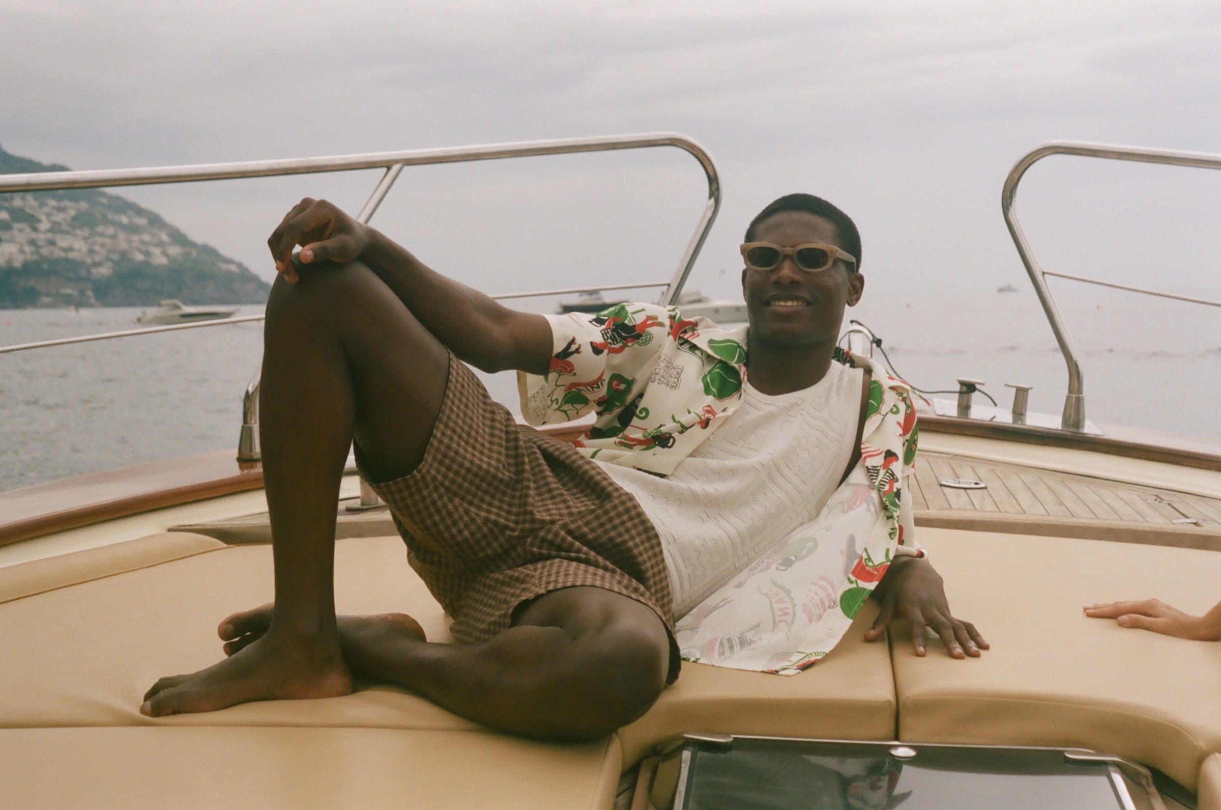 Man sitting on a boat with scenic background, wearing Le Sirenuse all-over patterned shirt. 