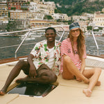 Two people sitting with a scenic view of a coastal town in the background, wearing Le Sirenuse x Tombolo pieces. 