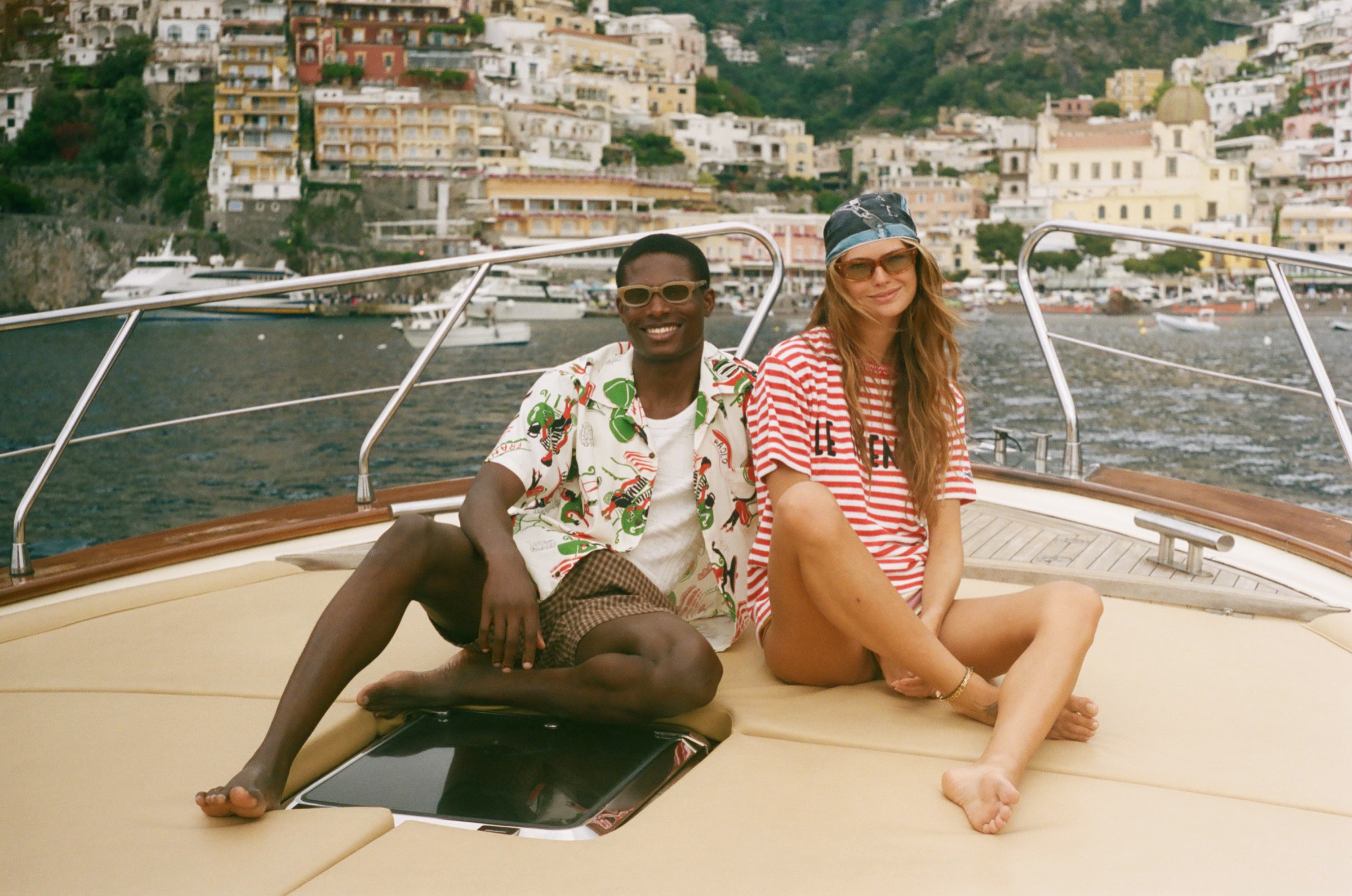 Two people sitting with a scenic view of a coastal town in the background, wearing Le Sirenuse x Tombolo pieces. 