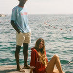 Two people on a dock by the water, one standing and one sitting, with a clear sky, both wearing peices from the Le Sirenuse x Tombolo collection.