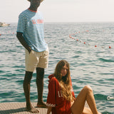 Two people on a dock by the water, one standing and one sitting, with a clear sky, both wearing peices from the Le Sirenuse x Tombolo collection.