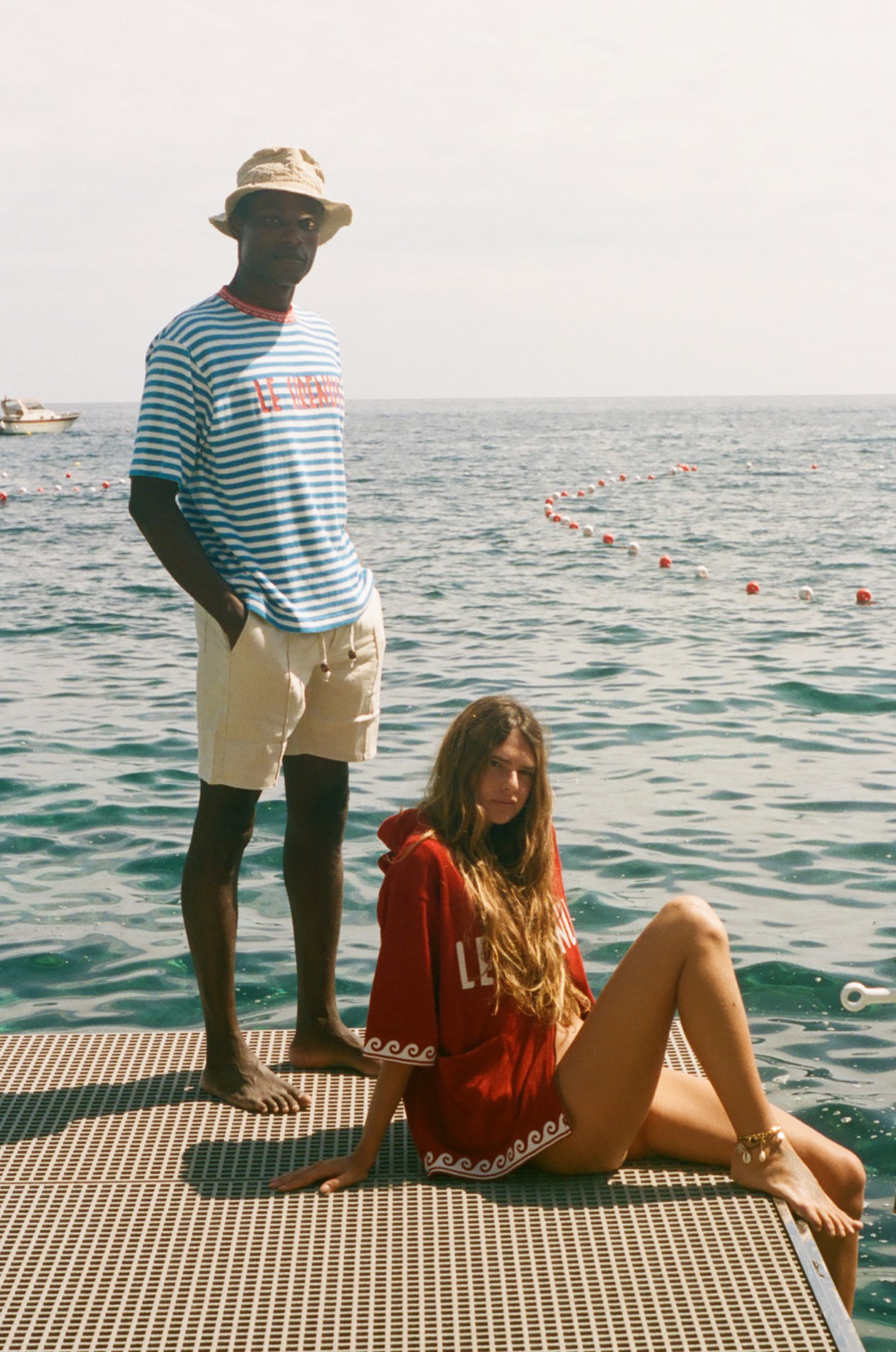 Two people on a dock by the water, one standing and one sitting, with a clear sky, both wearing peices from the Le Sirenuse x Tombolo collection.