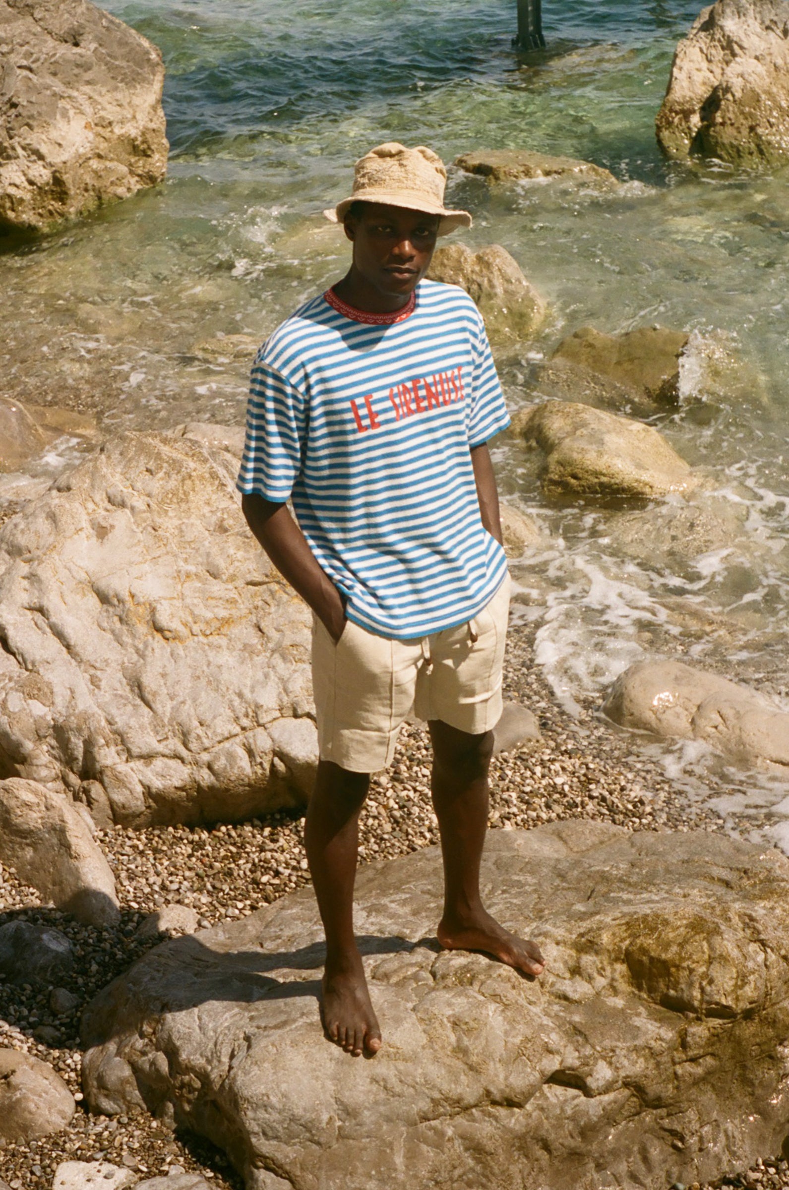 Man standing on rocks by the sea wearing a striped Le Sirenuse shirt.