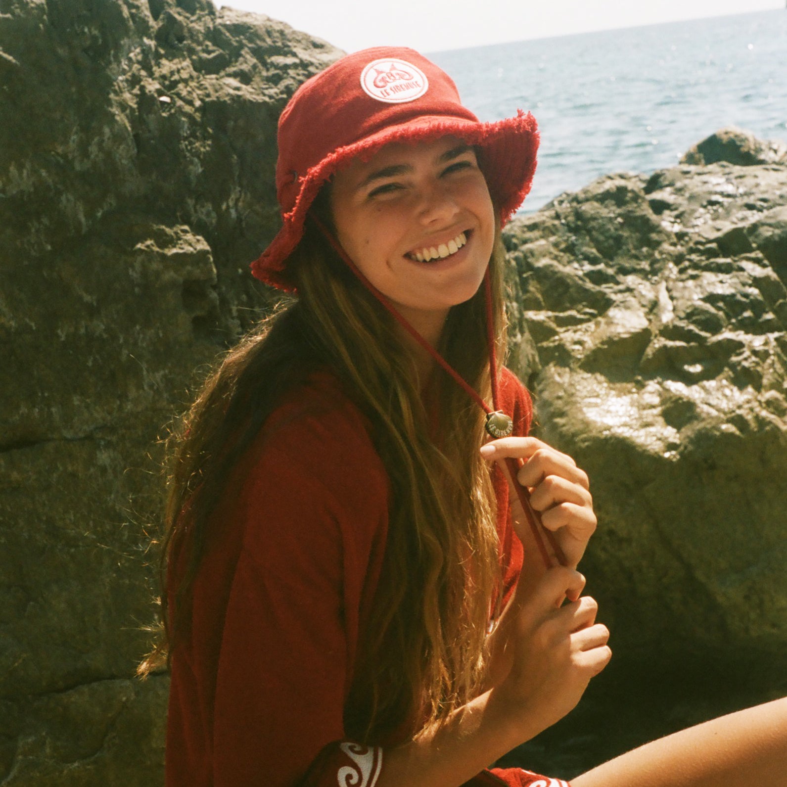 Woman wearing a red Le Sirenuse hat and outfit sitting on rocks by the ocean.