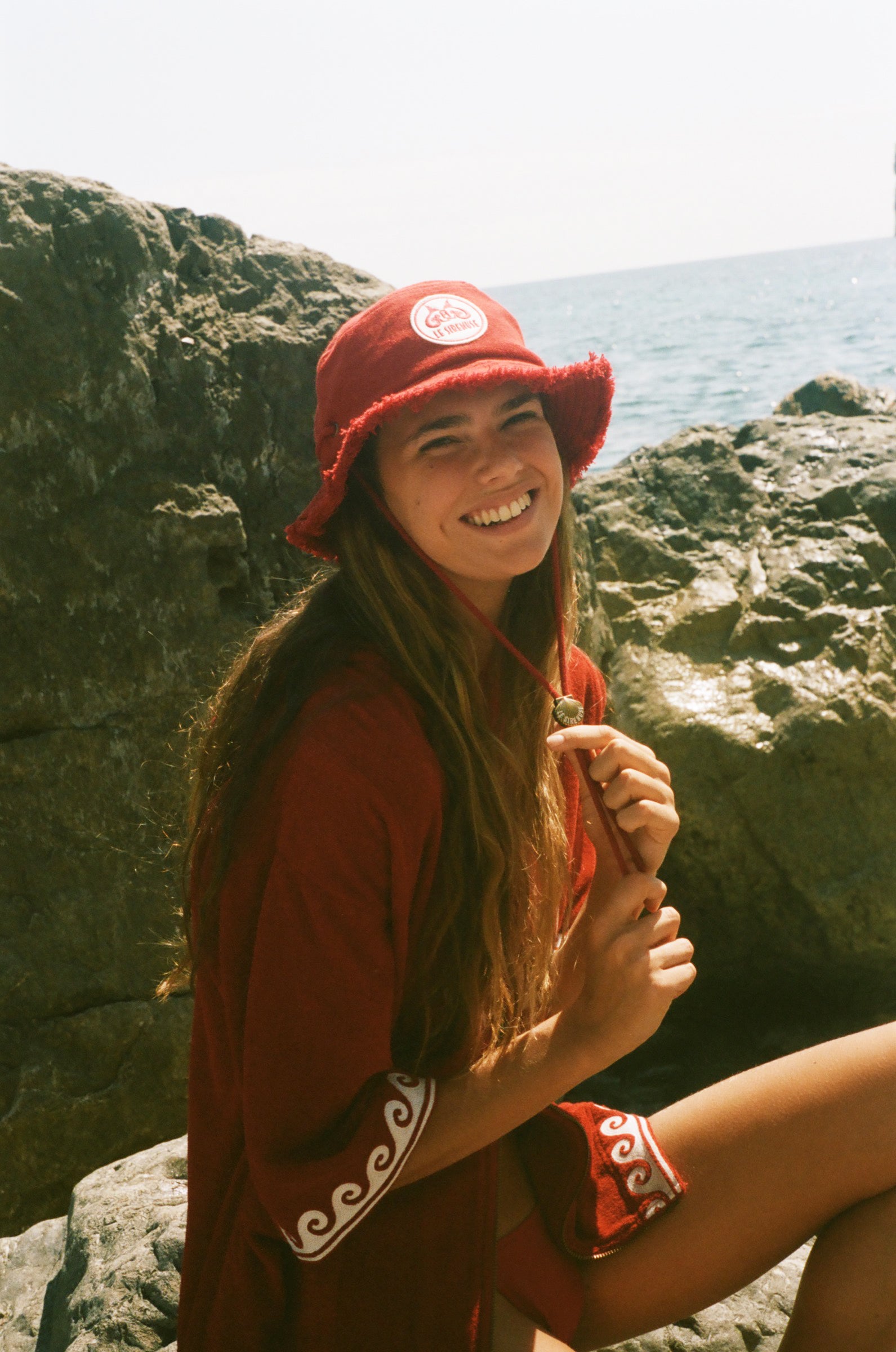 Woman wearing a red Le Sirenuse hat and outfit sitting on rocks by the ocean.