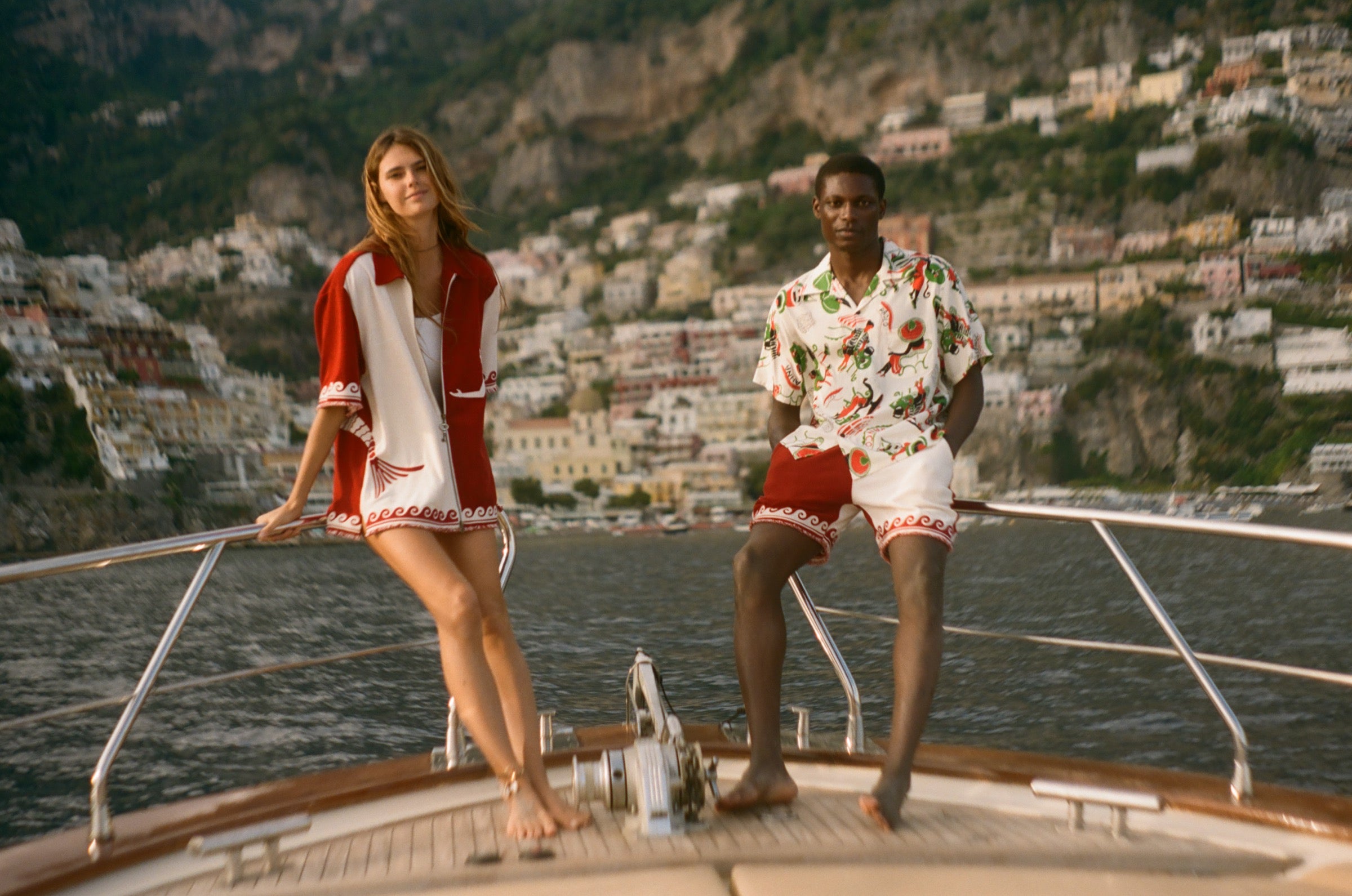 Two people on a boat with scenic mountains in the background, wearing items from the Le Sirenuse x Tombolo collection.