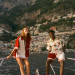 Two people standing on a boat with a scenic view of a coastal town and mountains in the background.
