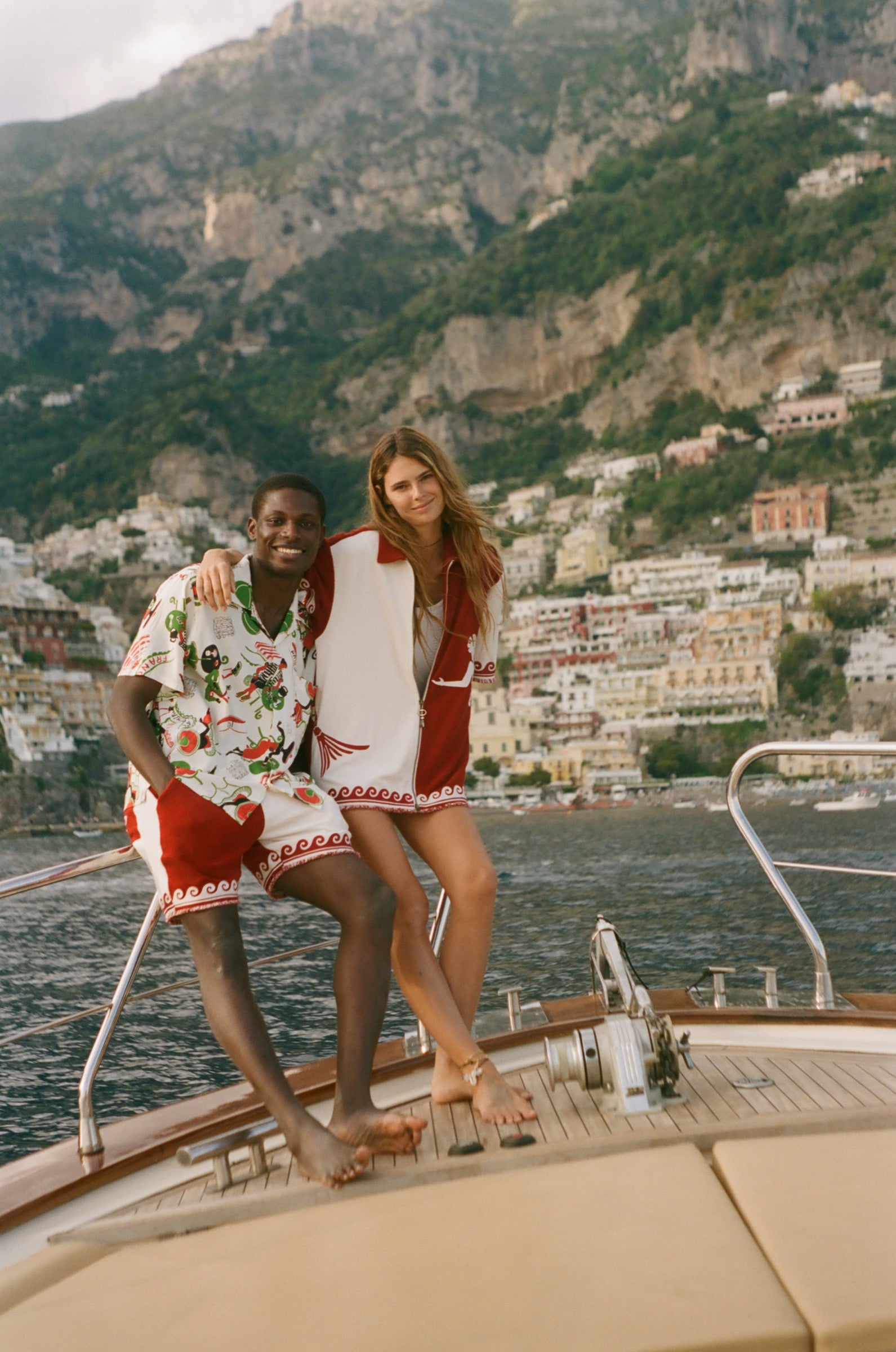 Two people standing on a boat with a scenic view of a coastal town and mountains in the background.