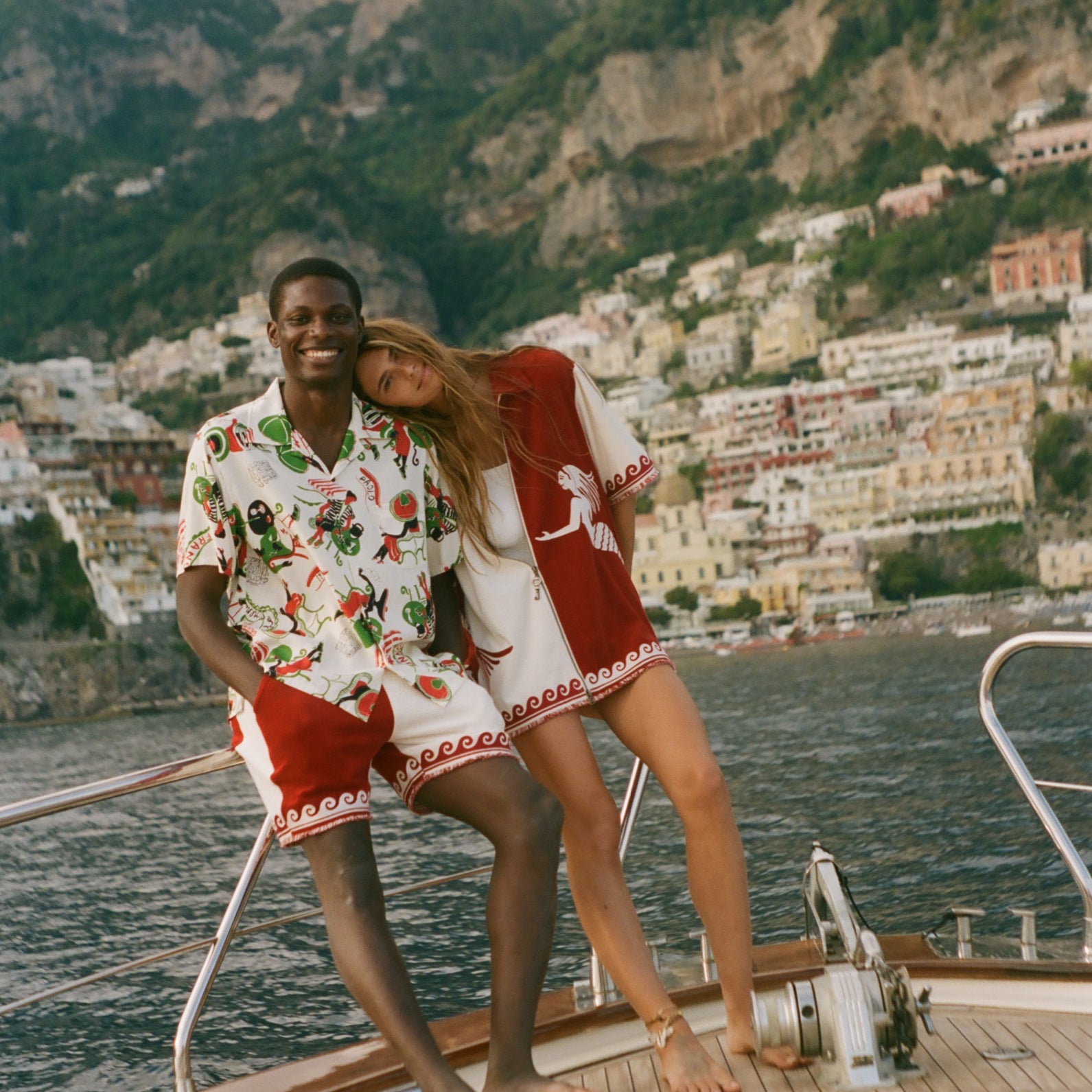 Two people on a boat with a scenic view of a coastal town and mountains, wearing Le Sirenuse x Tombolo collection pieces. 