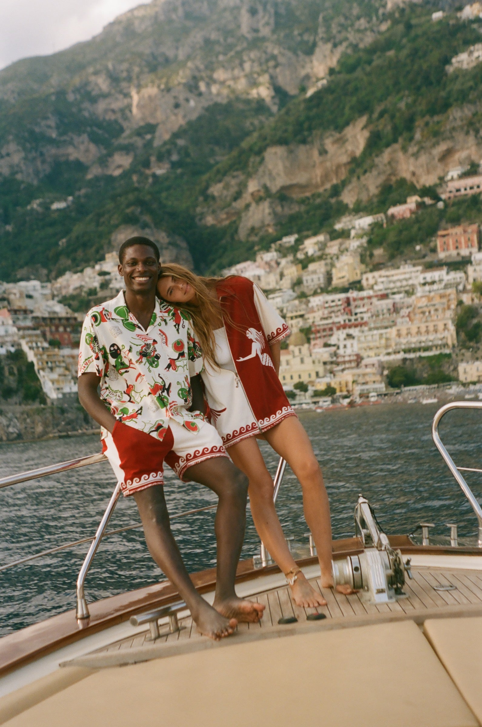 Two people on a boat with a scenic view of a coastal town and mountains, wearing Le Sirenuse x Tombolo collection pieces. 