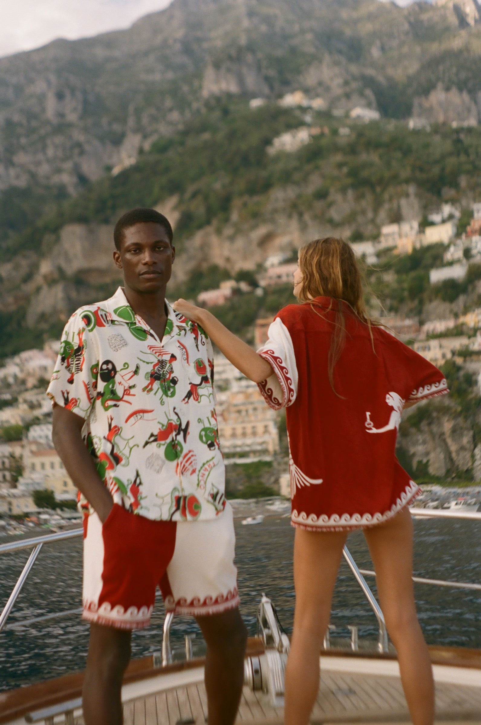 Two people standing on a boat with a scenic view of a coastal town and mountains in the background.