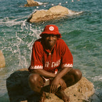 Person wearing a red Le Sirenuse shirt and hat sitting on a rock by the ocean