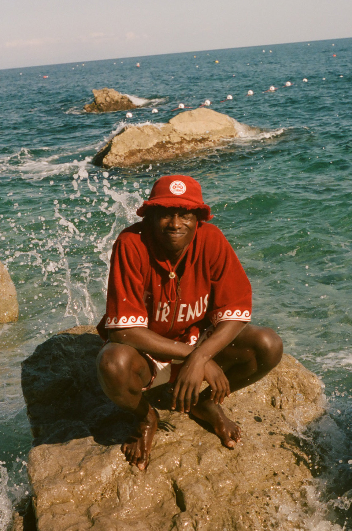 Person wearing a red Le Sirenuse shirt and hat sitting on a rock by the ocean