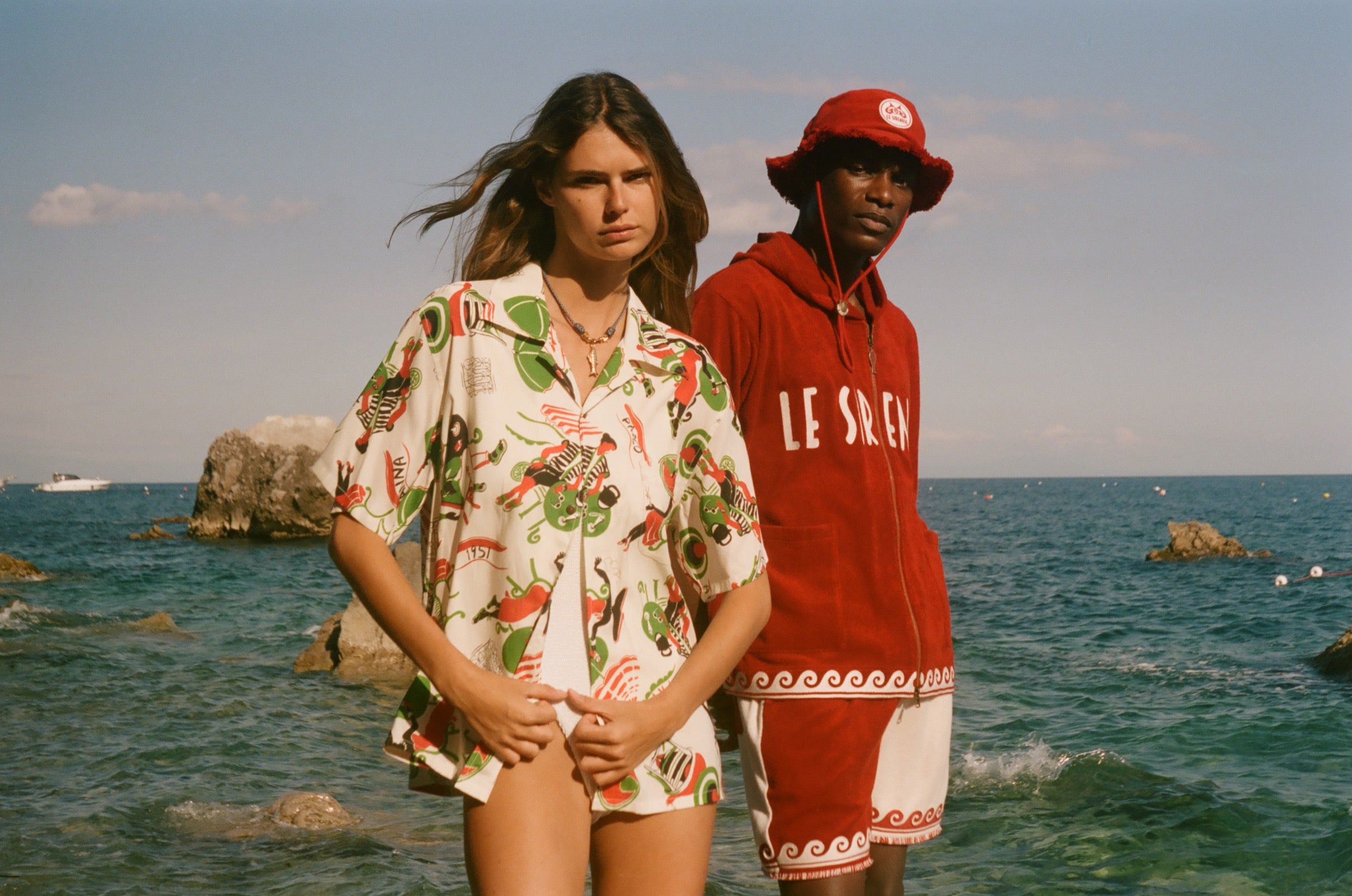 Two people standing in front of the beach, wearing Le Sirenuse x Tombolo collection items. 