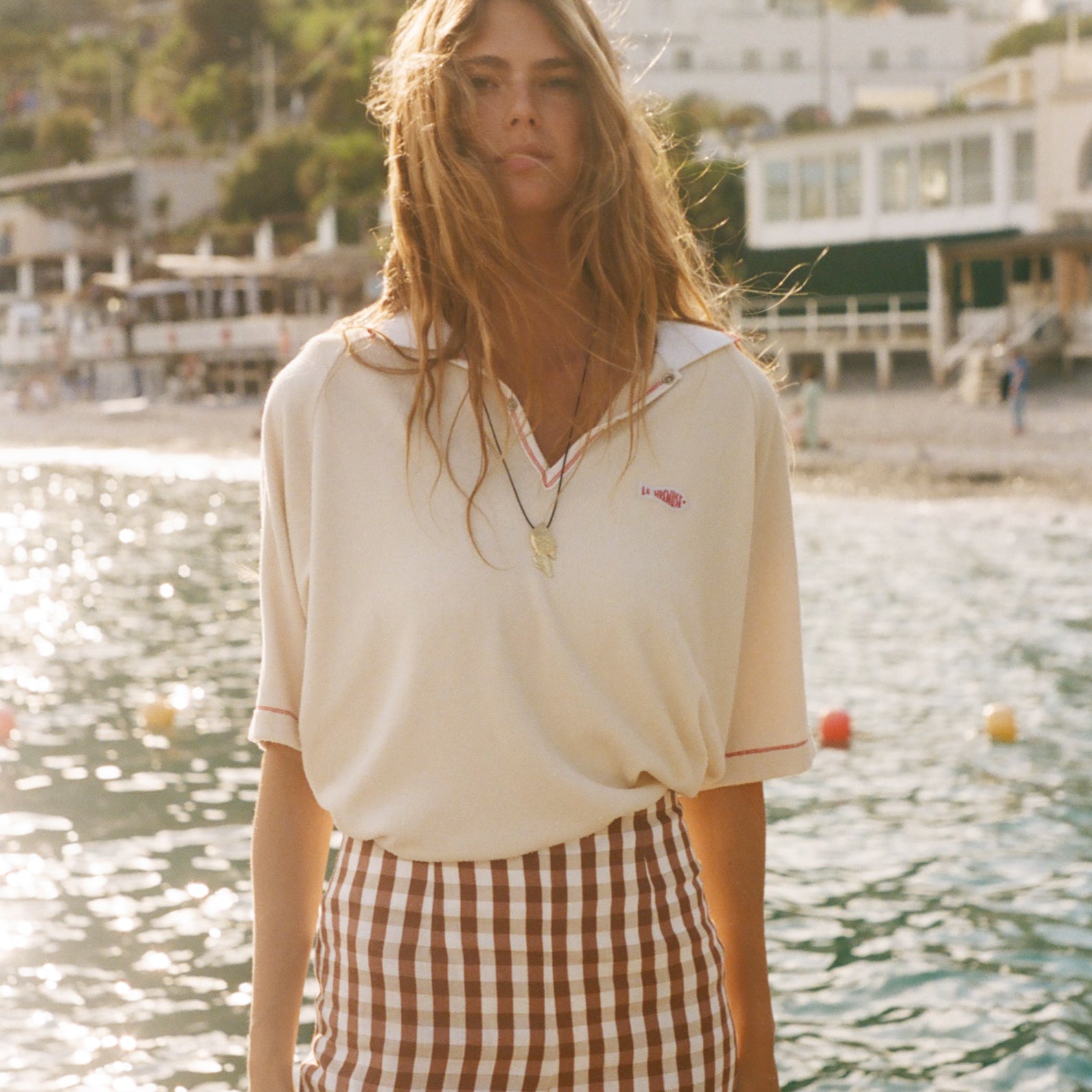 Woman standing by a waterfront, wearing the Le Sirenuse beige polo shirt. 