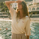 Woman standing by a waterfront with buildings and mountains in the background, wearing the beige Le Sirenuse polo shirt.