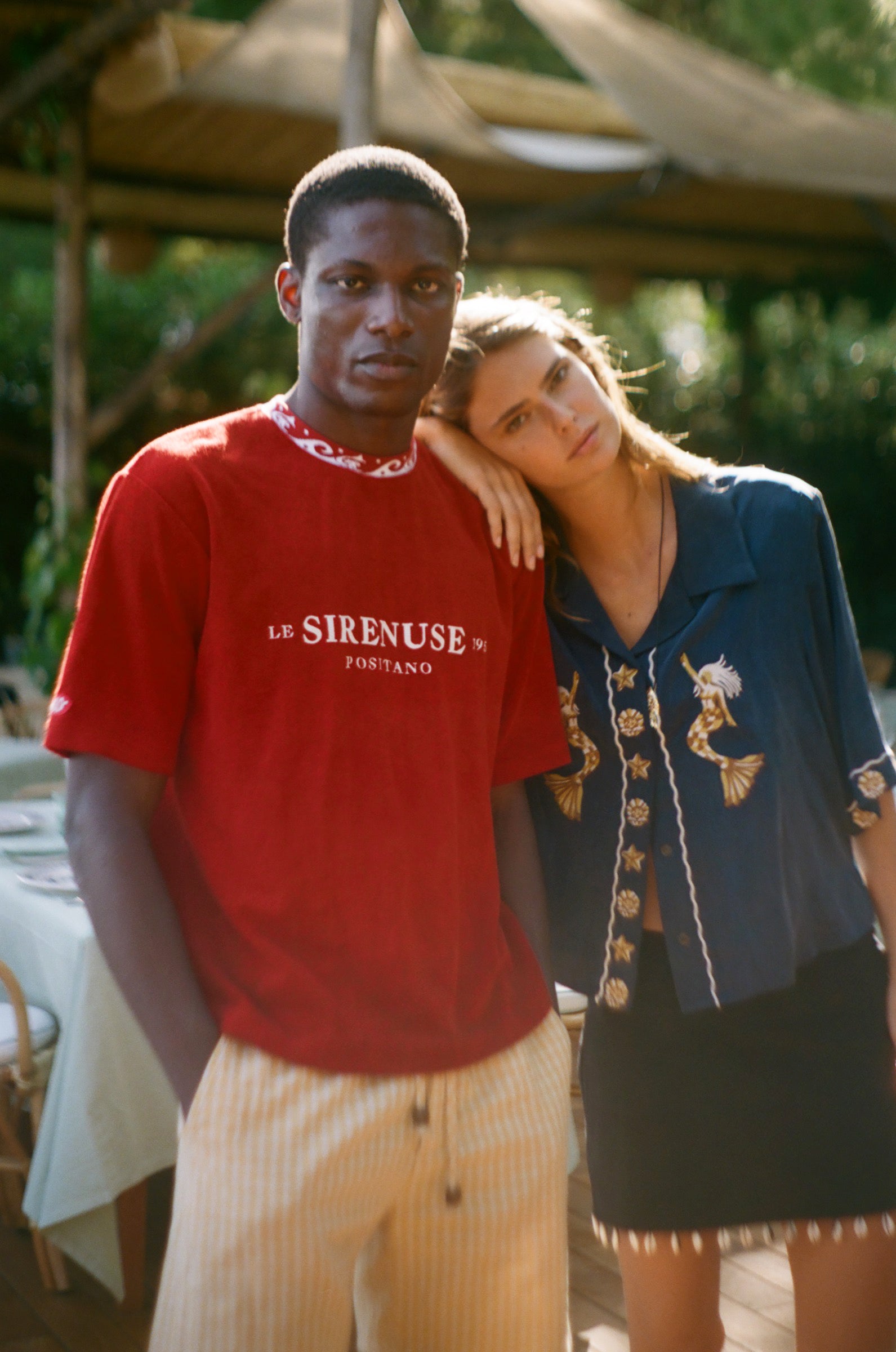 Two people standing close together outdoors with a red shirt featuring 'Le Sireneuse' branding.