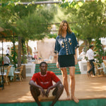 Woman standing on a wooden platform with a man sitting below, surrounded by greenery and people in the background.