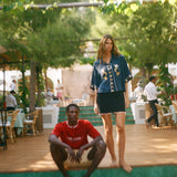 Woman standing on a wooden platform with a man sitting below, surrounded by greenery and people in the background.