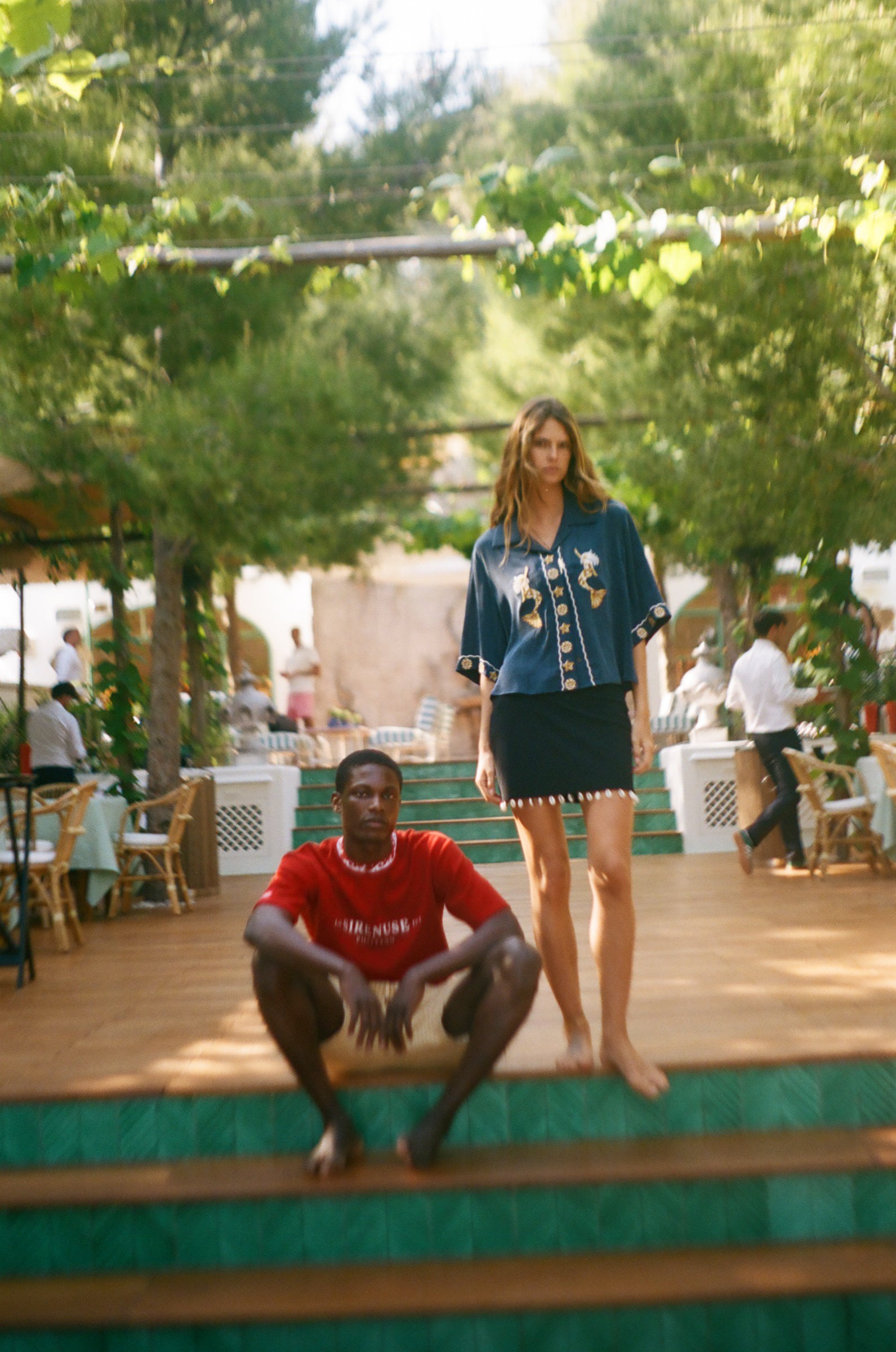 Woman standing on a wooden platform with a man sitting below, surrounded by greenery and people in the background.