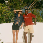 Two people standing on a sandy surface, wearing Le Sirenuse x Tombolo clothing with trees in the background
