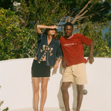 Two people standing on a sandy surface, wearing Le Sirenuse x Tombolo clothing with trees in the background