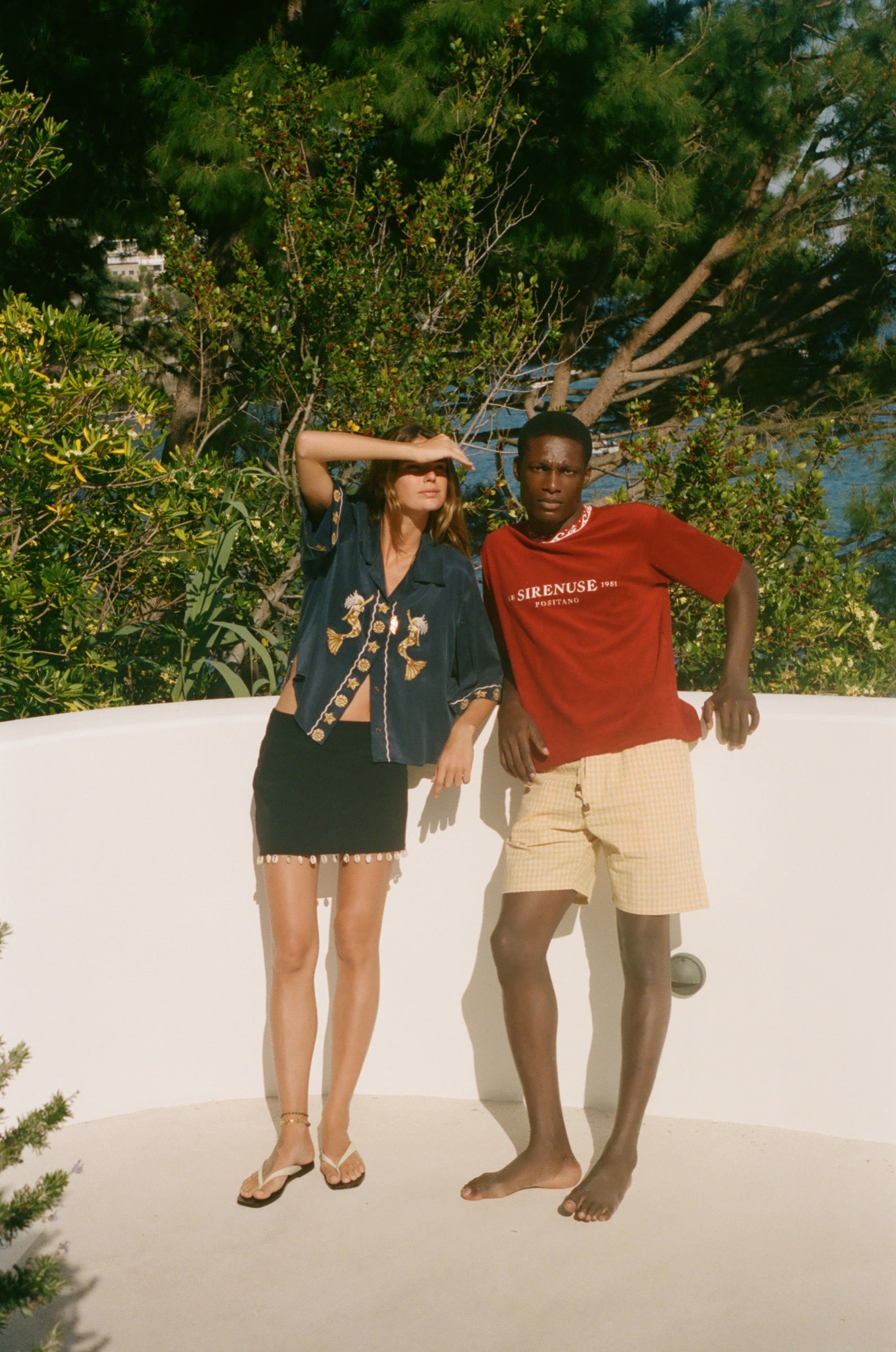 Two people standing on a sandy surface, wearing Le Sirenuse x Tombolo clothing with trees in the background