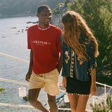 Two people standing on a balcony with a scenic view of water and mountains, wearing Le Sirenuse x Tombolo merchandise. 