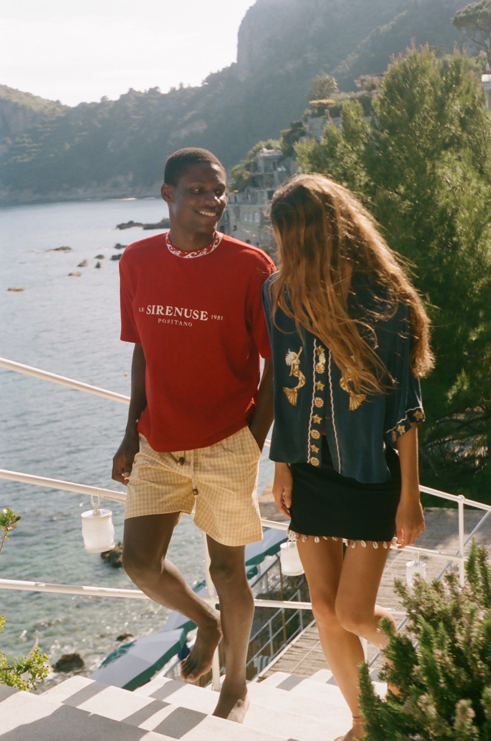 Two people standing on a balcony with a scenic view of water and mountains, wearing Le Sirenuse x Tombolo merchandise. 