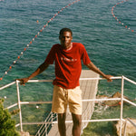 Man in a red shirt and beige shorts standing on a balcony overlooking the ocean with boats in the distance.