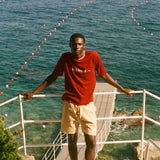Man in a red shirt and beige shorts standing on a balcony overlooking the ocean with boats in the distance.