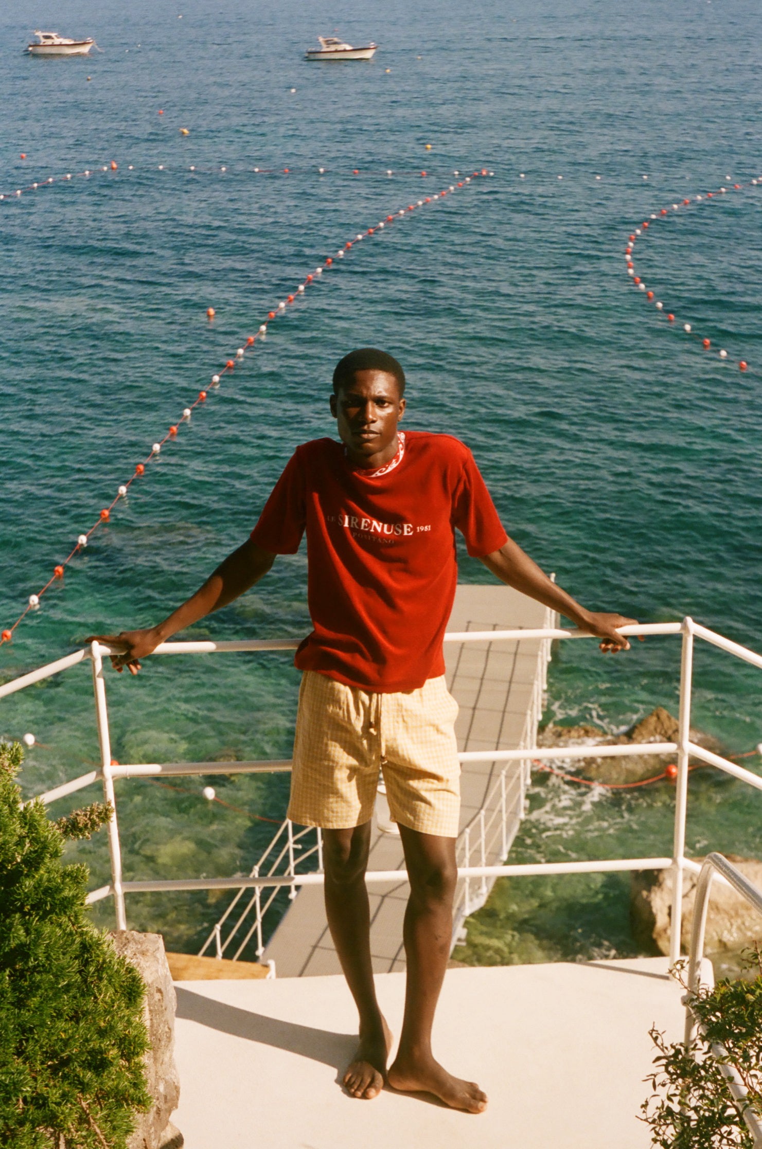 Man in a red shirt and beige shorts standing on a balcony overlooking the ocean with boats in the distance.