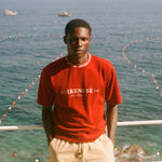 Man wearing Le Sirenuse red t-shirt, standing in front of the ocean. 