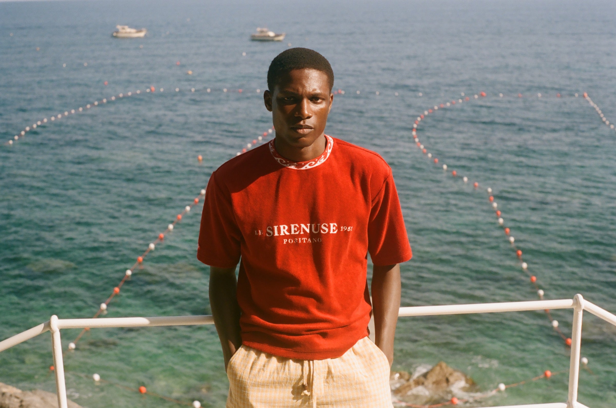 Man wearing Le Sirenuse red t-shirt, standing in front of the ocean. 