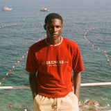 Man wearing Le Sirenuse red t-shirt, standing in front of the ocean. 