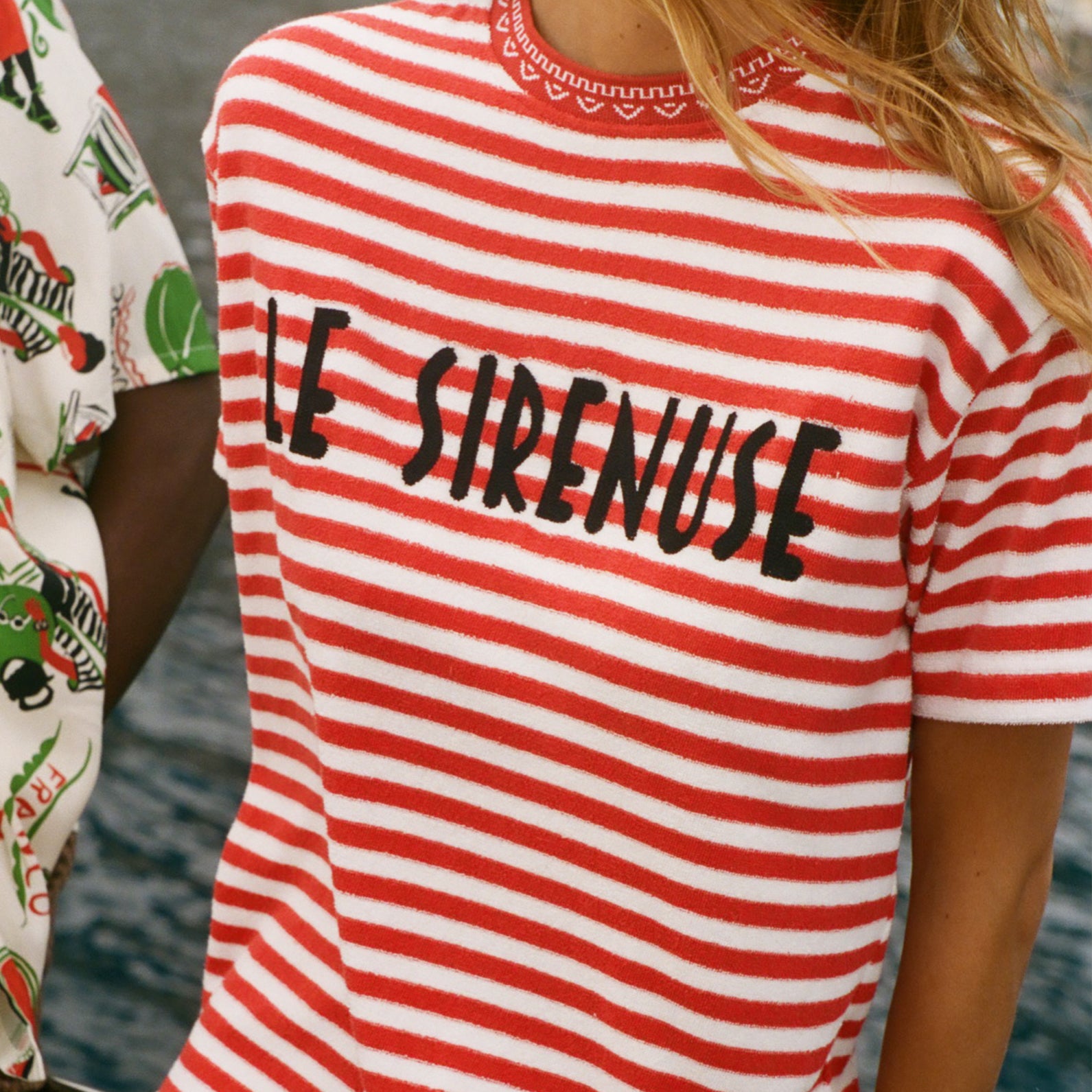 Person wearing a red and white striped shirt with 'LE SIRENUSE' text, standing by water.