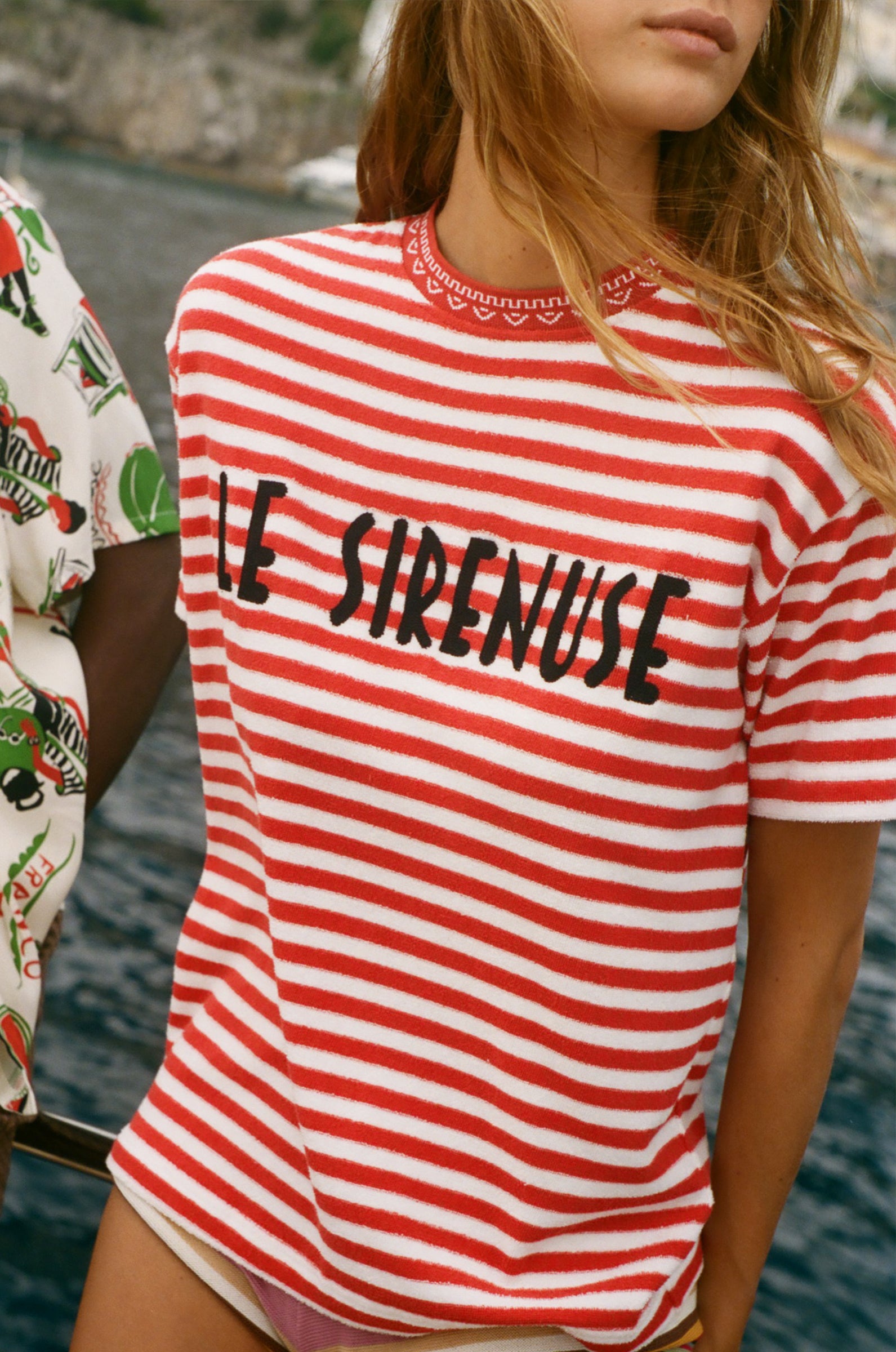 Person wearing a red and white striped shirt with 'LE SIRENUSE' text, standing by water.