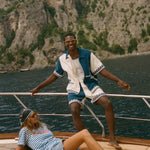 Two people on a sailboat with a scenic background, wearing items from the Le Sirenuse x Tombolo collection.