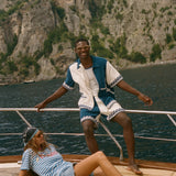 Two people on a sailboat with a scenic background, wearing items from the Le Sirenuse x Tombolo collection.