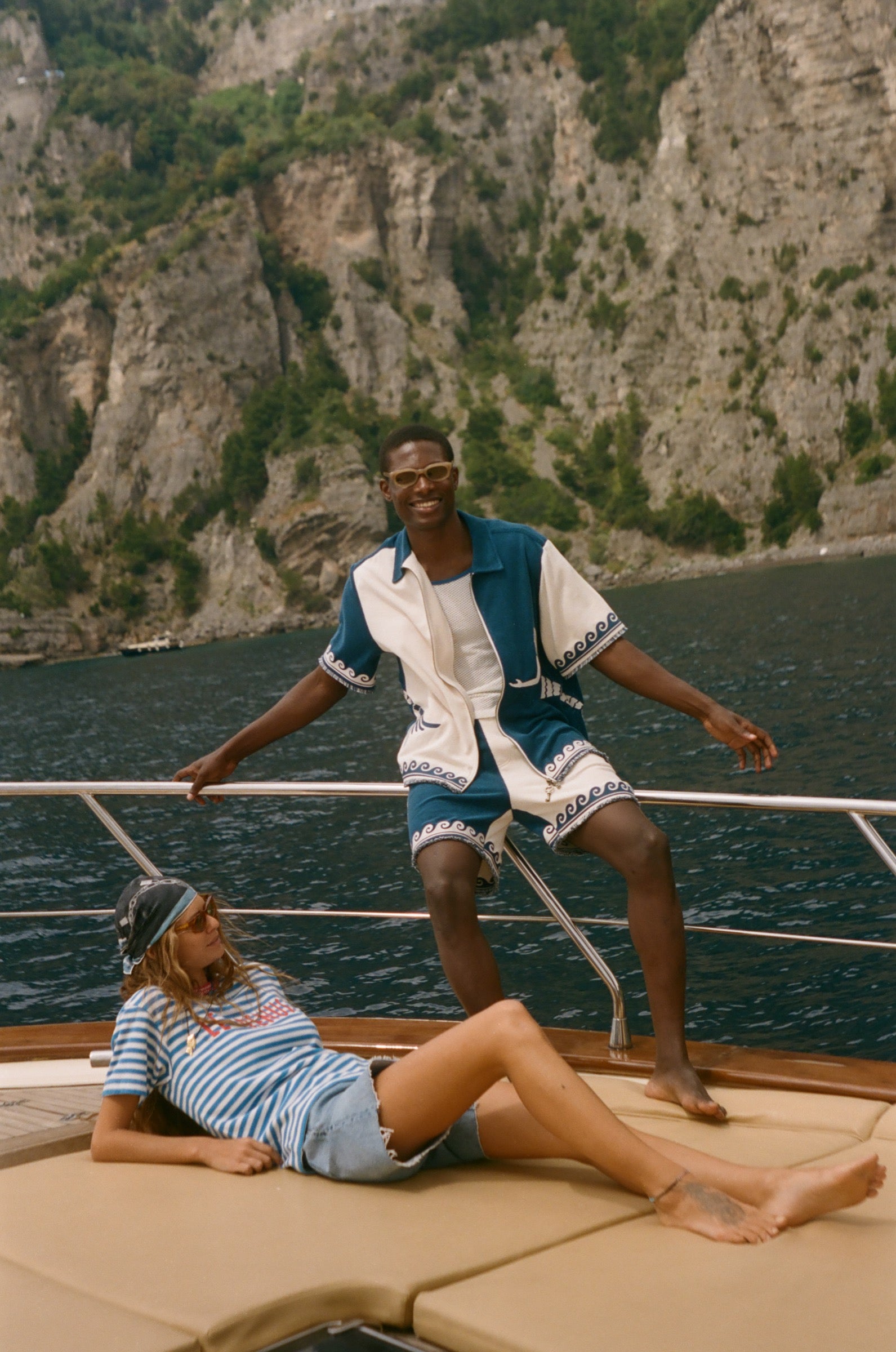 Two people on a sailboat with a scenic background, wearing items from the Le Sirenuse x Tombolo collection.