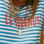 Person wearing a blue and white striped shirt with 'LE SIRENUSE' text, accessorized with necklaces.