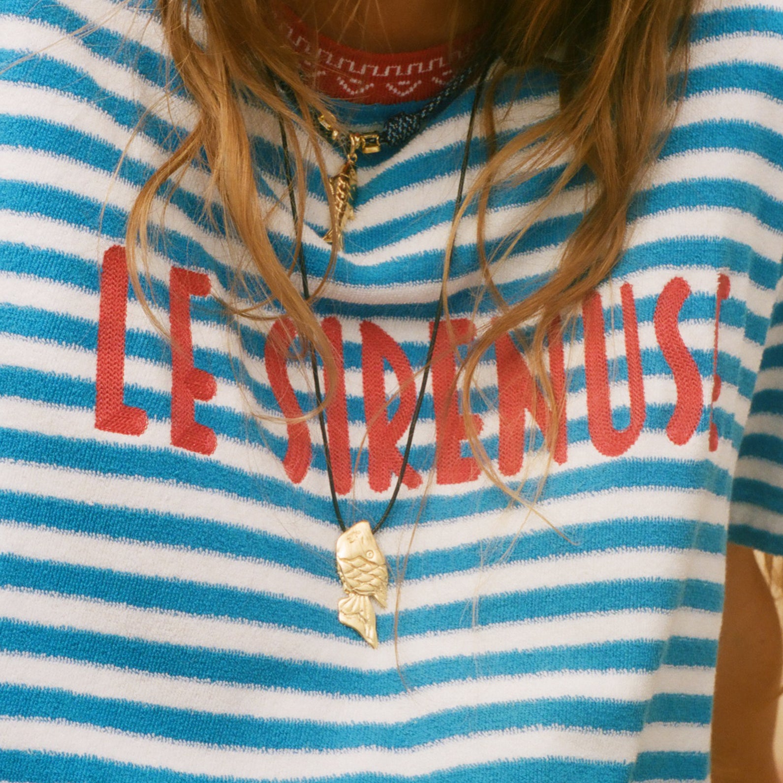 Person wearing a blue and white striped shirt with 'LE SIRENUSE' text, accessorized with necklaces.