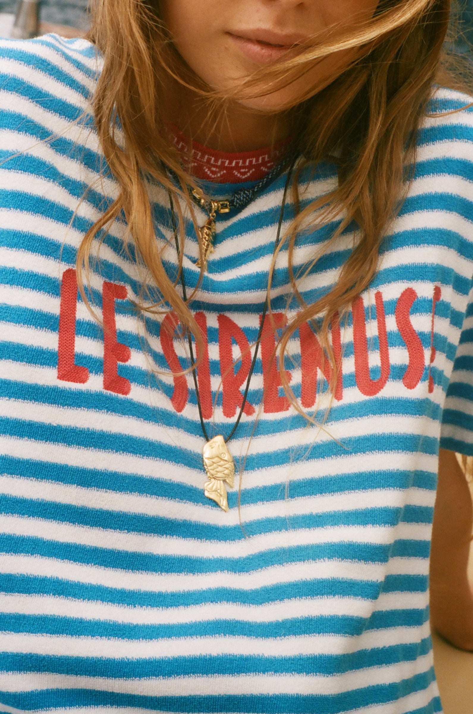 Person wearing a blue and white striped shirt with 'LE SIRENUSE' text, accessorized with necklaces.