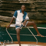 Man sitting on the edge of a boat, wearing the blue Late Checkout Cabana set. 