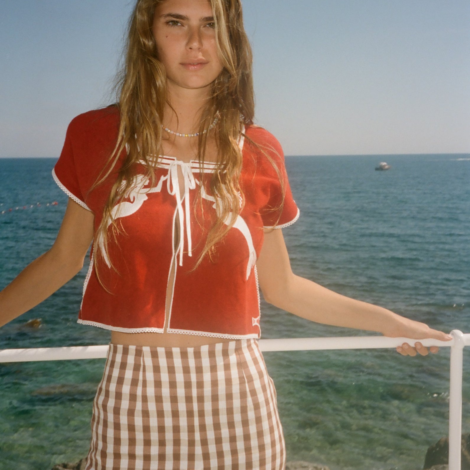 Woman in a red le sirenuse shirt and checkered pants standing on a boat with water in the background
