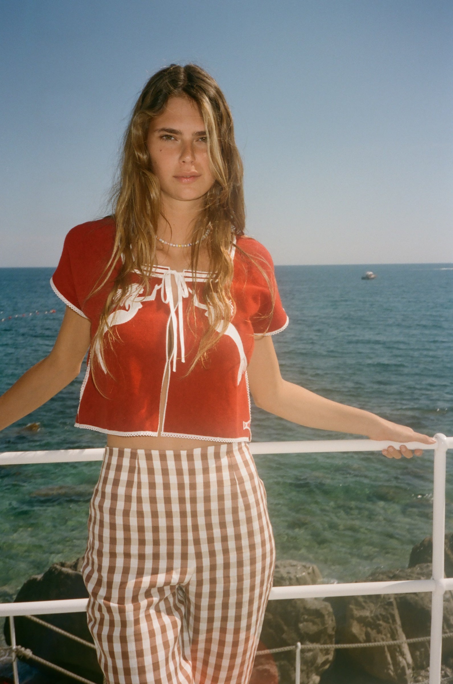Woman in a red le sirenuse shirt and checkered pants standing on a boat with water in the background