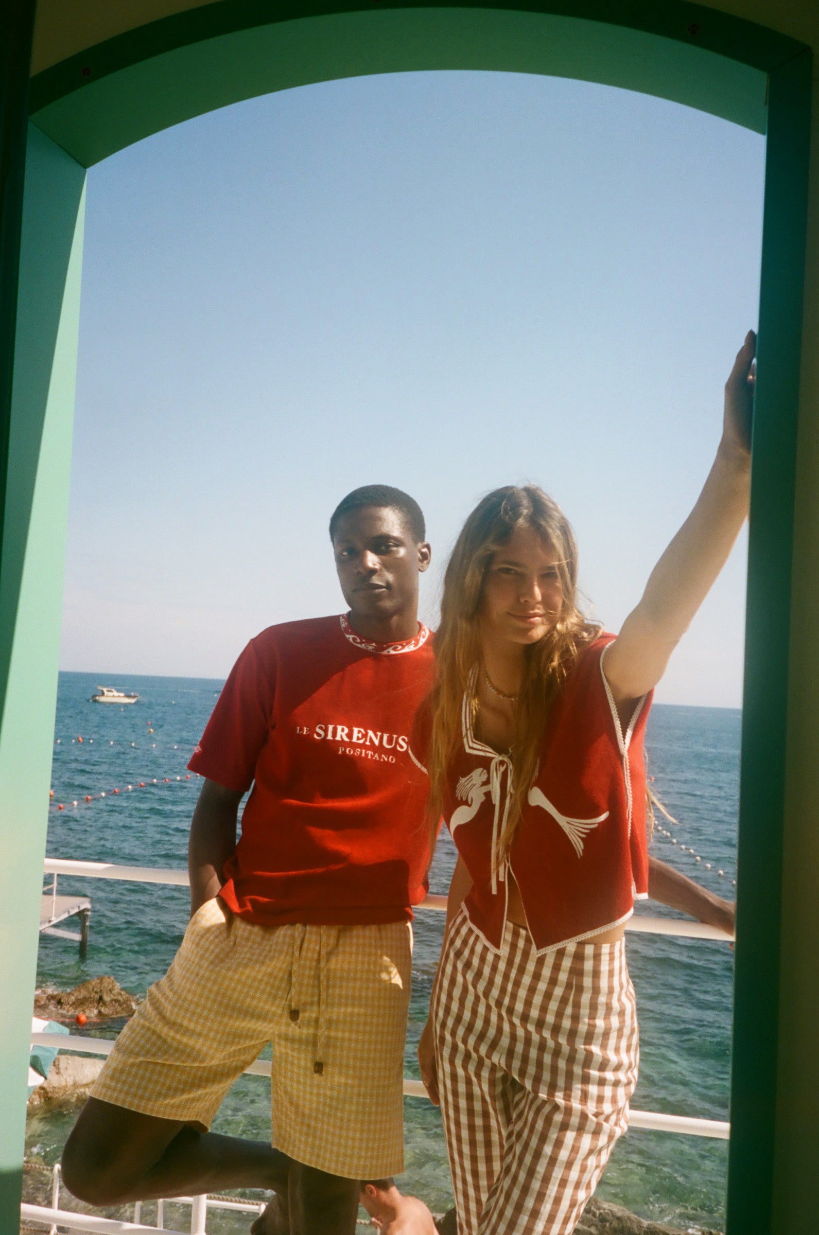 Two people standing by a scenic ocean view with a green frame, wearing shirts from the Le Sirenuse x Tombolo collaboration.